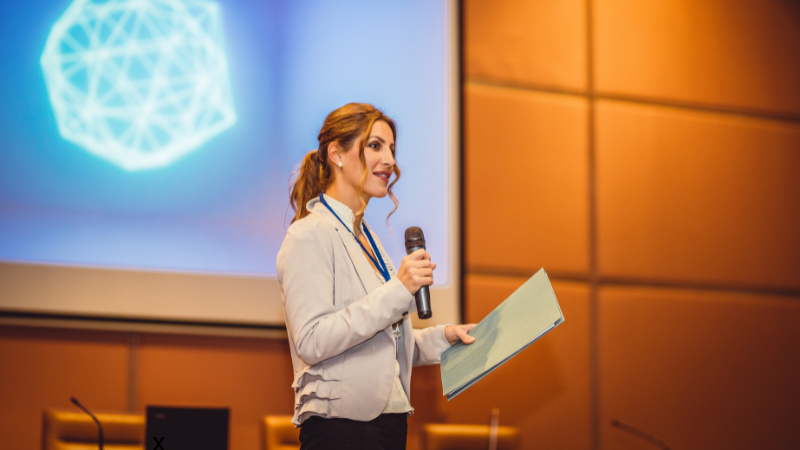 woman holding a microphone giving a presentation in front of an audience